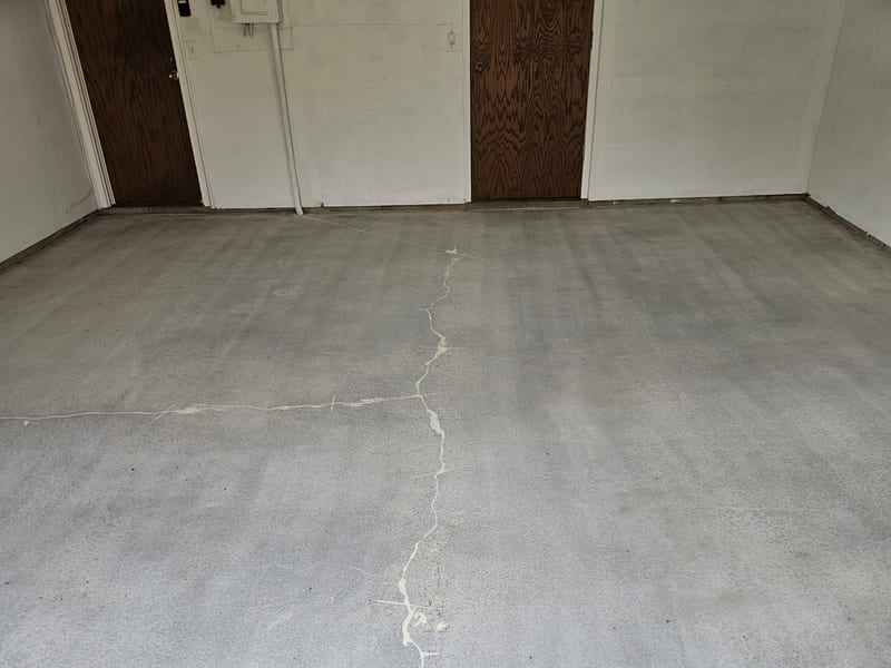 The same Wayzata garage floor diamond-ground to bare grey concrete, with the full-depth crack routed out and filled with white repair compound down the length of the slab